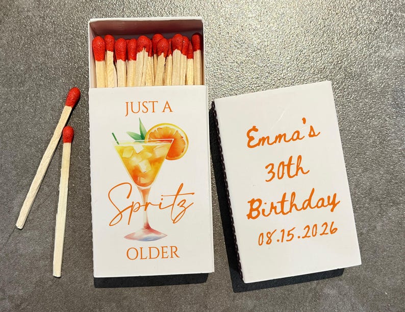May include: A white matchbox with the text "JUST A Spritz OLDER" and a cocktail illustration. The open box reveals red-tipped matches. The adjacent lid reads "Emma's 30th Birthday 08.15.2026" in orange.