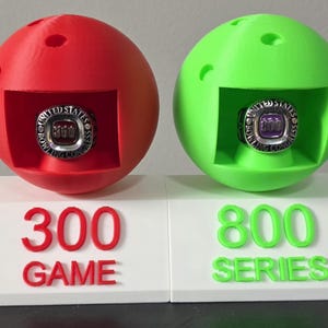 May include: A red and a green bowling ball-shaped display stand, each holding a ring. The red stand has the text "300 GAME", and the green stand has "800 SERIES". The rings are silver with purple and white accents.