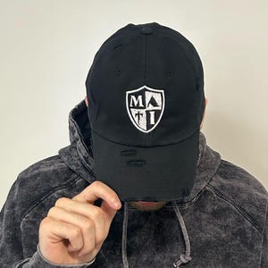May include: Black baseball cap with a distressed brim. The cap features a white shield-shaped emblem with the letters "MA" and "I". The person in the image is wearing a black and gray hoodie.