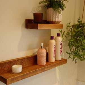 May include: Two wooden shelves mounted on a white wall. The top shelf has a glass candle holder and a plant in a white pot. The bottom shelf has three bottles of lotion and a white candle.