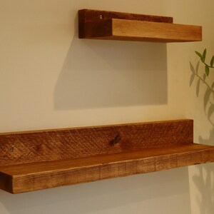 May include: Two wooden shelves mounted on a white wall. The top shelf is smaller and the bottom shelf is longer. Both shelves are made of reclaimed wood and have a natural brown finish.