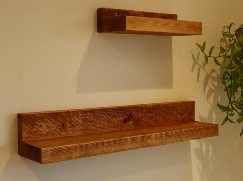 May include: Two wooden shelves mounted on a white wall. The top shelf is smaller and the bottom shelf is longer. Both shelves are made of reclaimed wood and have a natural brown finish.
