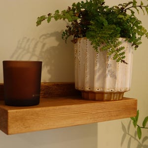 May include: A wooden shelf with a brown finish, mounted on a wall. A brown glass candle and a white ceramic planter with a green plant are on the shelf.