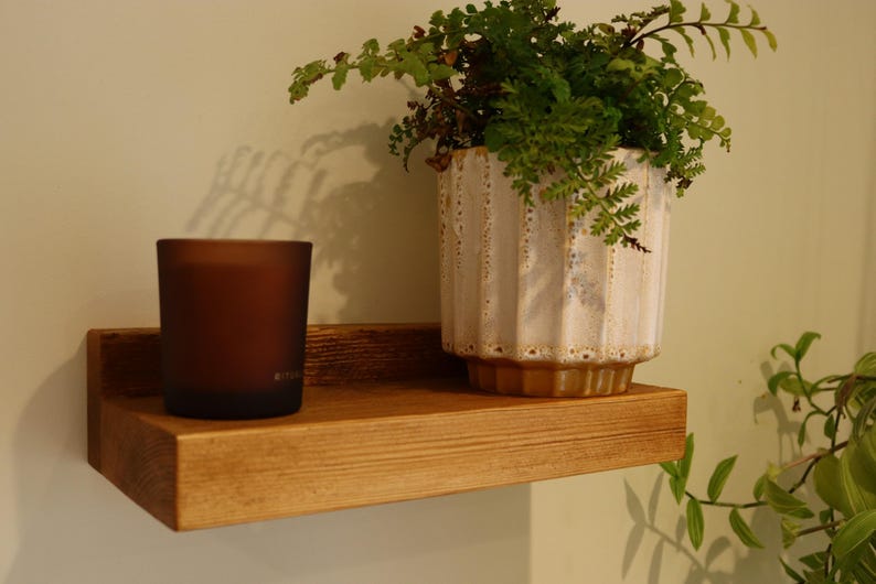 May include: A wooden shelf with a brown finish, mounted on a wall. A brown glass candle and a white ceramic planter with a green plant are on the shelf.
