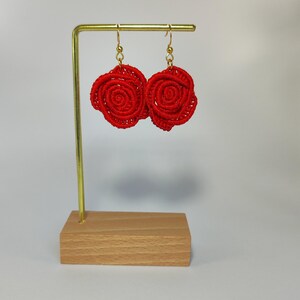 May include: A pair of red rose-shaped earrings with gold-colored hooks. The earrings are displayed on a gold-colored metal stand with a wooden base. The earrings are made of a woven material.