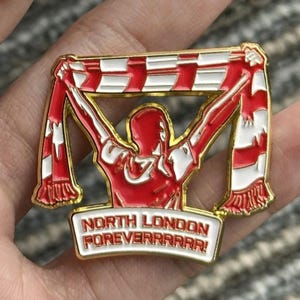 May include: A red and white enamel pin with a gold border. The pin features a figure holding up a scarf with a red and white pattern. The text "NORTH LONDON FOREVERRRRR!" is displayed on a white banner.