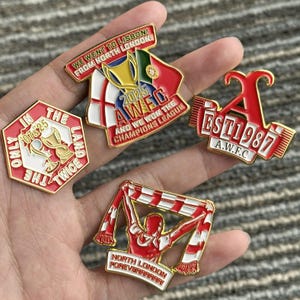 Puede incluir: Un conjunto de pines esmaltados rojos, blancos y dorados. Uno de ellos presenta el texto "WE WENT TO LISBON! FROM NORTH LONDON!" y un trofeo. Otro dice "EST 1987 A.W.F.C.". Los pines probablemente están relacionados con un equipo deportivo.