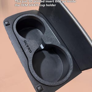 May include: A black 3D-printed cup holder insert for the OEM GX470 cup holder. The insert features two circular cup holders and the text "GX470". The insert is designed to fit inside the original cup holder.