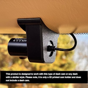 May include: A black 3D printed dash cam holder designed to fit a dash cam with a similar style. The holder is mounted to the windshield of a car. The text "FOCUM RE CEO RoHS" is visible on the dash cam.