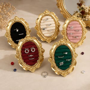 Peut inclure: Cinq présentoirs à bagues ovales avec des cadres dorés ornés. Chaque présentoir possède un intérieur doublé de velours noir, crème, rose, rouge foncé ou vert foncé, avec des fentes horizontales pour les bagues. Plusieurs bagues argentées sont insérées.