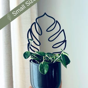 Puede incluir: Un pequeño soporte para plantas de metal negro con forma de hoja de monstera. El soporte sostiene una pequeña planta en maceta con hojas verdes.