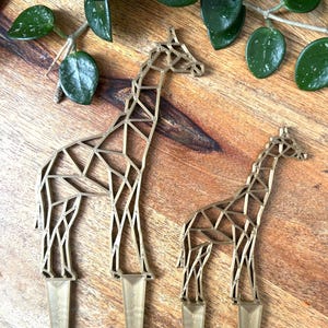 May include: Two gold geometric giraffe cake toppers. The larger giraffe is standing and the smaller giraffe is standing with its head turned to the side. Both giraffes have a pointed base.