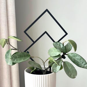 Puede incluir: Una maceta de cerámica blanca con nervaduras verticales, que contiene una planta verde con hojas verde plateado. Un enrejado de plantas de metal negro en forma de diseño geométrico se encuentra detrás de la planta.