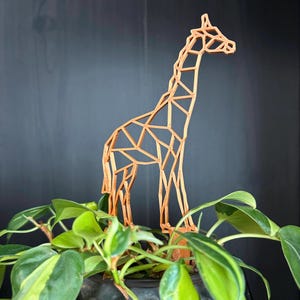 May include: A wooden geometric giraffe sculpture in a natural wood colour. The giraffe is standing in a potted plant with green leaves.