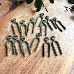 以下が含まれることがあります： 木製の表面に配置されたオリーブグリーンの植物クリップのコレクション。各クリップはU字型で、縞模様のトップが付いています。背景には緑の葉と茎が見えます。