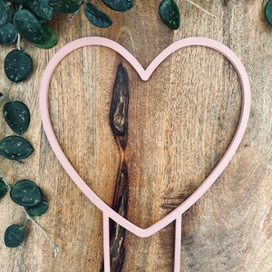 May include: A pink metal heart-shaped plant stand with two long stems. The stand is on a wooden surface.