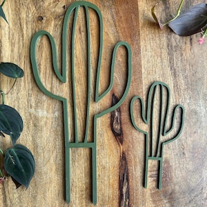 May include: Two green wooden cactus cutouts on a wood background. The larger cactus is taller and has a single arm, while the smaller cactus is shorter and has two arms.