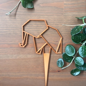May include: A copper-colored geometric elephant cake topper. The elephant is made of a series of connected triangles and is standing on a thin, pointed base.
