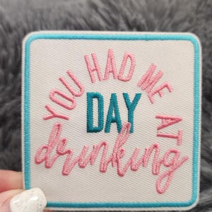 Op de afbeelding: Geborduurde patch met de tekst "YOU HAD ME AT DRINKING" in roze en teal garen. De patch heeft een witte achtergrond met een teal rand en ligt op een grijze, fuzzy ondergrond.