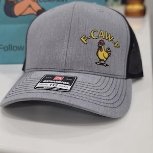 May include: A grey and black trucker hat featuring a yellow cartoon rooster graphic with the text F-CAW-F embroidered in yellow above it. The brim has a black sticker with the text RICHARDSON 112 TRUCKER EST 1970 ORIGINAL ADJUSTABLE.