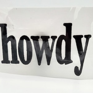 May include: Black "howdy" sticker. The word "howdy" is written in a bold, sans-serif font. The sticker is on a clear background.