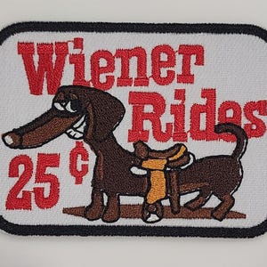 May include: Embroidered patch with a white background and a black border. The patch features a cartoon dachshund dog with a saddle, the text "Wiener Rides" in red, and the price "25¢". The dog is brown with a long snout and a smiling expression.