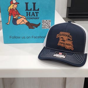 May include: A navy blue and white trucker hat with a leather patch that says "LOUISIANA PIPELINER". A blue shopping bag with the text "LL HAT COMPANY" and a cartoon image of a woman in a cowboy hat and boots.