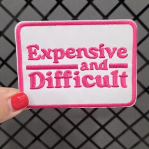Op de afbeelding: Een rechthoekige geborduurde patch met een witte achtergrond en een dikke roze rand. De tekst "Expensive and Difficult" is in roze draad in het midden geborduurd, met een horizontale roze lijn die de twee woorden van elkaar scheidt.