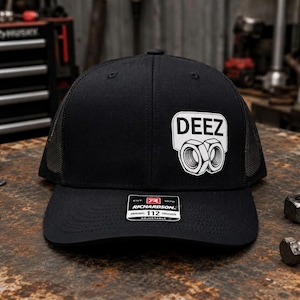 Könnte beinhalten: Eine schwarze Trucker-Cap mit Netzrücken und verstellbarem Riemen. Auf der Vorderseite befindet sich ein weißer Aufnäher mit dem Text DEEZ über einer Abbildung von zwei Sechskantmuttern aus Metall. Ein schwarz-weißer Aufkleber zeigt RICHARDSON 112 TRUCKER.