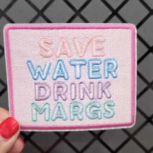 May include: A rectangular embroidered patch with a light pink background and a dark pink border. The text SAVE WATER DRINK MARGS is stitched in four rows using different thread colors: pink, blue, purple, and teal, in a rounded sans-serif font.