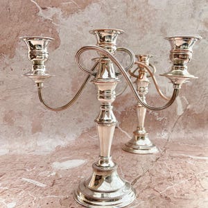 May include: A polished silver-toned candelabra featuring a central pillar and two curved arms, each topped with a candle cup. The metal surface is reflective and smooth, accented with beaded detailing along the base and the rims of the candle holders.