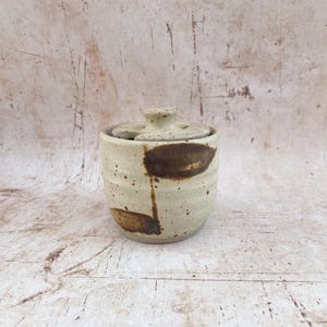 May include: A small, handmade ceramic jar with a lid. The jar is a light beige colour with brown abstract markings. The lid has a small knob for easy removal. The jar is likely used for storing small items or as a decorative piece.
