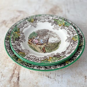 May include: Two stacked ceramic plates with a green rim and a brown and green grapevine border pattern. The centre of the top plate features a colourful illustration of two people sitting in a field with a dog, rendered in muted tones.