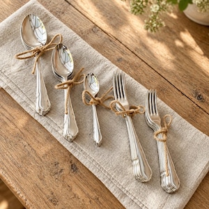 May include: A set of five bundles of silver-toned metal cutlery, including spoons and forks of varying sizes, each tied with a thin brown jute twine. The cutlery features a classic handle design and is arranged on a folded light beige linen napkin.