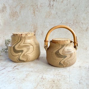 May include: Two beige ceramic jars with cork lids and a wavy brown design. One jar has a wooden handle. These jars are likely for storage or decorative purposes.