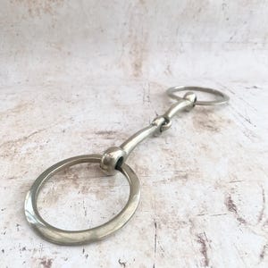 May include: A silver-coloured metal horse bit featuring two large circular rings connected by a central jointed bar. The equestrian equipment has a smooth, polished finish and a hinged design that allows for movement between the two side rings.