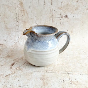May include: A ceramic jug with a rounded base and a spout. The jug has a light blue and white glaze, with a handle. The rim has a darker, brownish colour. This handmade jug is a decorative and functional kitchenware item, perfect for serving.