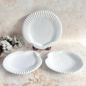 May include: A collection of three white ceramic plates, each shaped like a scallop shell. One large plate is displayed on a wooden stand, with two smaller plates below. The plates have a ribbed pattern, creating a seaside aesthetic.