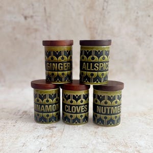 May include: A set of five cylindrical ceramic spice jars with dark brown wooden lids. Each jar features a repeating black and olive green geometric pattern and is labelled with the text GINGER, ALLSPICE, CINNAMON, CLOVES, or NUTMEG in black.