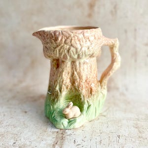 May include: A ceramic jug with a tree trunk design. The jug is mainly tan and brown, with a textured surface. A small, light-coloured rabbit figurine sits in a patch of green grass at the base.