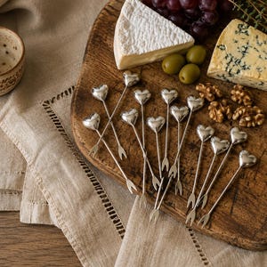 May include: A collection of silver-toned metal appetiser picks, each featuring a small heart-shaped topper and a two-pronged fork end. These slender utensils are arranged in a cluster, showcasing their polished finish and uniform design.
