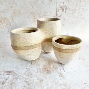 May include: A set of three connected ceramic vessels with a speckled cream and beige finish. The structure features two smaller rounded cups joined to a taller central cylindrical container, all sharing a consistent textured, earthy appearance.