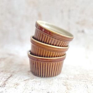 May include: Four brown ceramic ramekins are stacked. Each ramekin features a ribbed design on the outside and a light-coloured interior. The ramekins are arranged slightly off-centre. The colour is a warm, earthy brown, suitable for baking and serving.