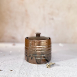 May include: A handcrafted, brown ceramic jar with a lid. The jar has a cylindrical shape with horizontal ridges and dark, dripped accents. The lid has a small knob. The jar is likely used for storage or decoration.