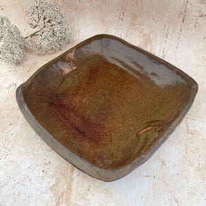 May include: A square ceramic dish with a brown glaze. The dish has raised edges and a textured surface. The overall aesthetic is rustic and artisanal. Ideal for serving appetizers or small dishes. The dish is handmade.