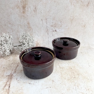 May include: Two dark brown ceramic lidded bowls. The bowls are round with a small knob on top of the lid. The bowls are on a white and beige surface. The bowls are likely for cooking or serving.