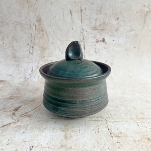 May include: A teal ceramic jar with a lid, featuring a dark brown rim and base. The lid has a small, rounded handle. The jar's body has horizontal grooves. The background is a light, textured surface. The jar is likely handmade.