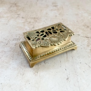 May include: A rectangular brass box featuring an ornate, openwork lid decorated with embossed floral and leaf motifs. The base of the box is elevated on four small feet and is detailed with a beaded border along the lower edge of the container.
