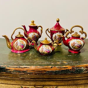 May include: Five decorative miniature teapots and a sugar bowl. The teapots are a deep red with gold accents and handles. Each teapot features a painted scene of people in a garden. The sugar bowl has a matching design.