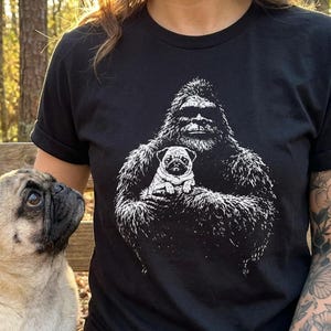Könnte beinhalten: Schwarzes T-Shirt mit einem weißen Aufdruck von Bigfoot, der einen Mops hält. Ein echter Mops-Hund sitzt auf einer Holzbank neben der Person, die das Shirt trägt. Der Hintergrund ist ein sonniger Wald.
